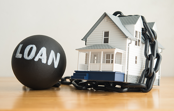 a dollhouse on a hardwood floor with a ball and chain attached to it; the ball has the word loan written in white paint